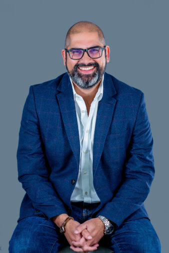 A man with a beard and glasses, wearing a blue blazer, white shirt, and jeans, sits smiling against a plain blue background.
