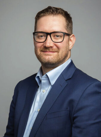 A man with short brown hair, beard, and glasses, wearing a blue suit jacket and light blue shirt, poses in front of a plain gray background.