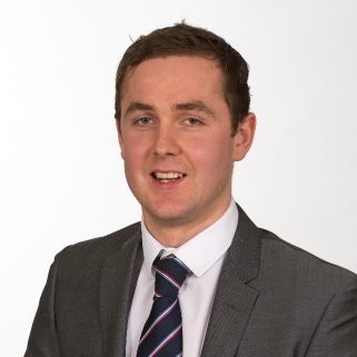 A man in a grey suit, white shirt, and striped tie poses for a professional headshot against a plain white background.