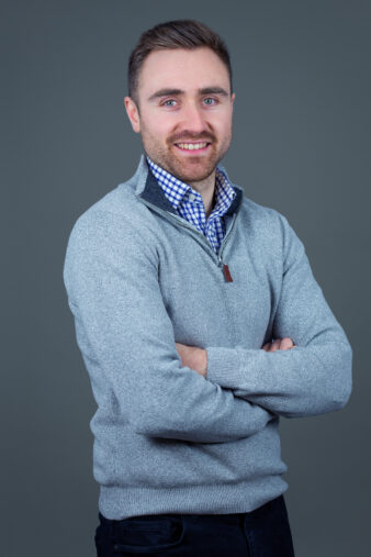 A man with short brown hair and a beard stands with arms crossed, wearing a light gray sweater over a checkered shirt, in front of a plain gray background.