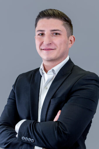 A man in a black suit and white shirt stands with arms crossed, smiling slightly, in front of a plain gray background.