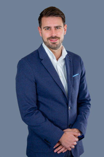 A man with brown hair and a beard, wearing a blue suit and white shirt, stands with his hands folded against a plain gray background.