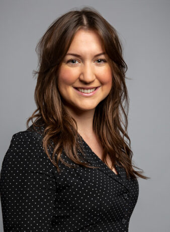 A woman with long brown hair smiles at the camera. She is wearing a black blouse with white polka dots and stands against a plain gray background.