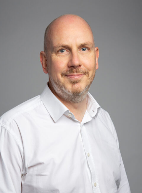 A middle-aged man with a shaved head and short beard, wearing a white dress shirt, poses against a plain gray background, smiling slightly at the camera.