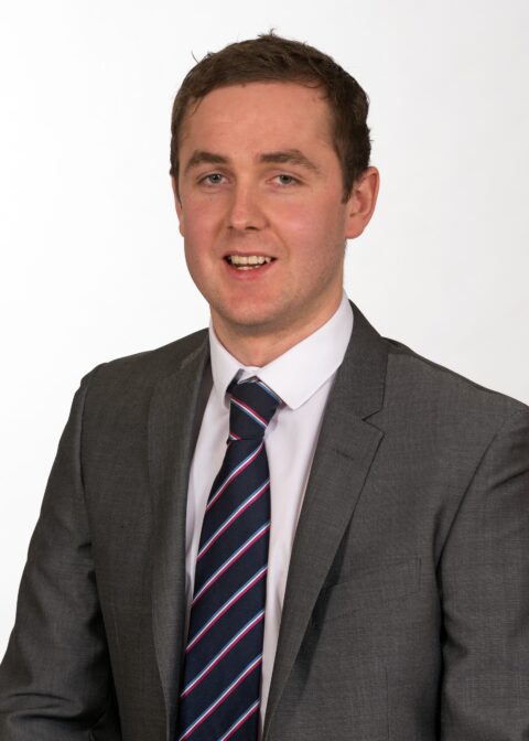 A man in a grey suit, white shirt, and striped tie posed against a plain white background.