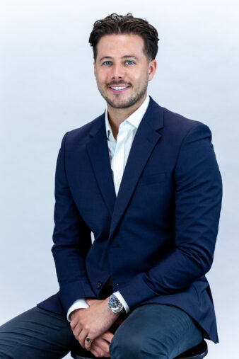 A man with short dark hair and a beard, wearing a navy blazer, white shirt, and dark trousers, sits on a stool against a plain light background.