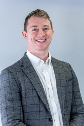 A man in a gray plaid suit jacket and white shirt stands smiling against a light gray background.