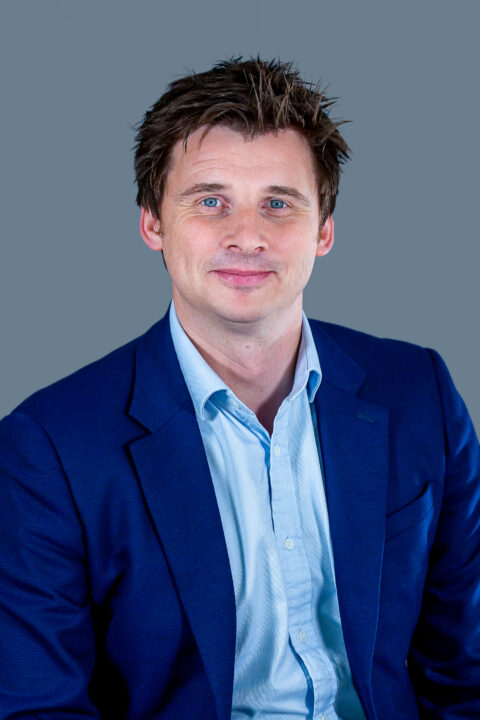A man with short brown hair wearing a blue suit jacket and light blue shirt sits against a plain grey background, smiling slightly.