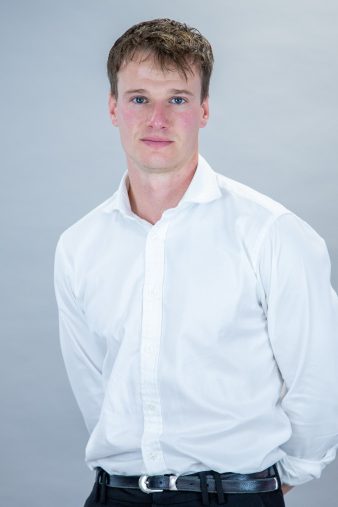 Person wearing a white dress shirt and black trousers, standing against a plain light grey background, looking directly at the camera with a neutral expression.