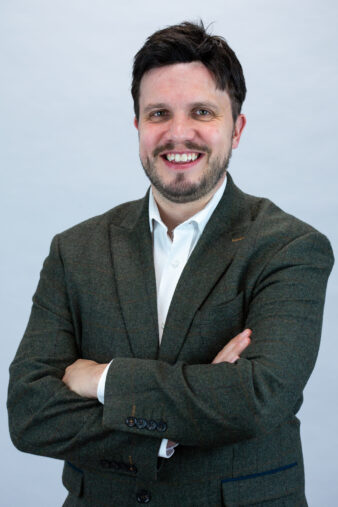 Man with dark hair and beard, wearing a white shirt and green blazer, stands with arms folded and smiles at the camera against a plain light background.