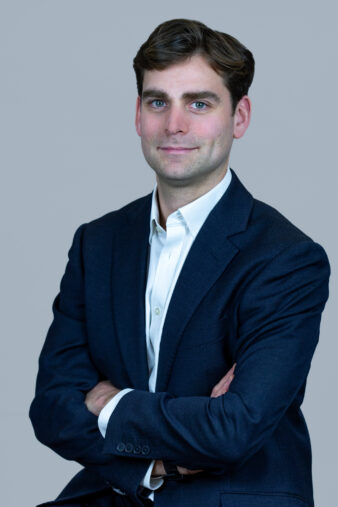 A man with brown hair in a dark suit and white shirt poses against a plain gray background with his arms crossed.
