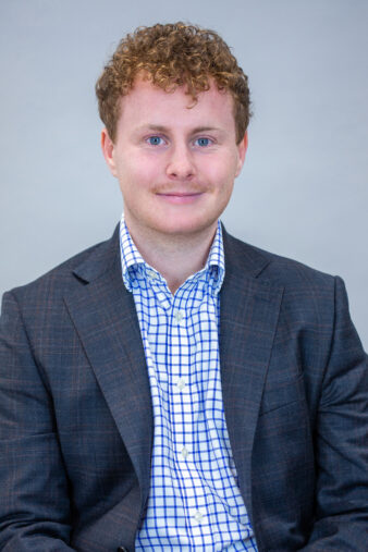 A man with curly light brown hair, wearing a dark suit jacket and a blue and white checkered shirt, sits facing the camera against a plain light background.