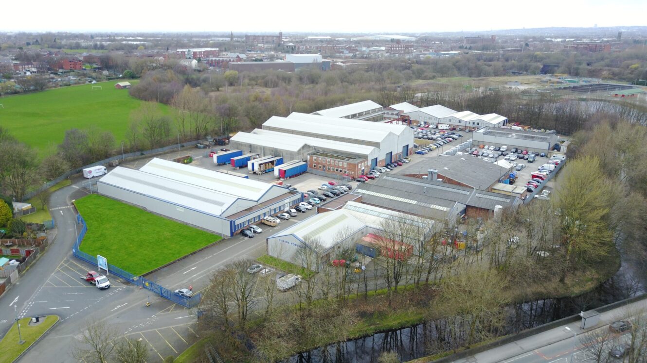 Aerial view of an industrial complex with multiple warehouses, parking lots filled with vehicles, and surrounding trees and roads on a cloudy day.