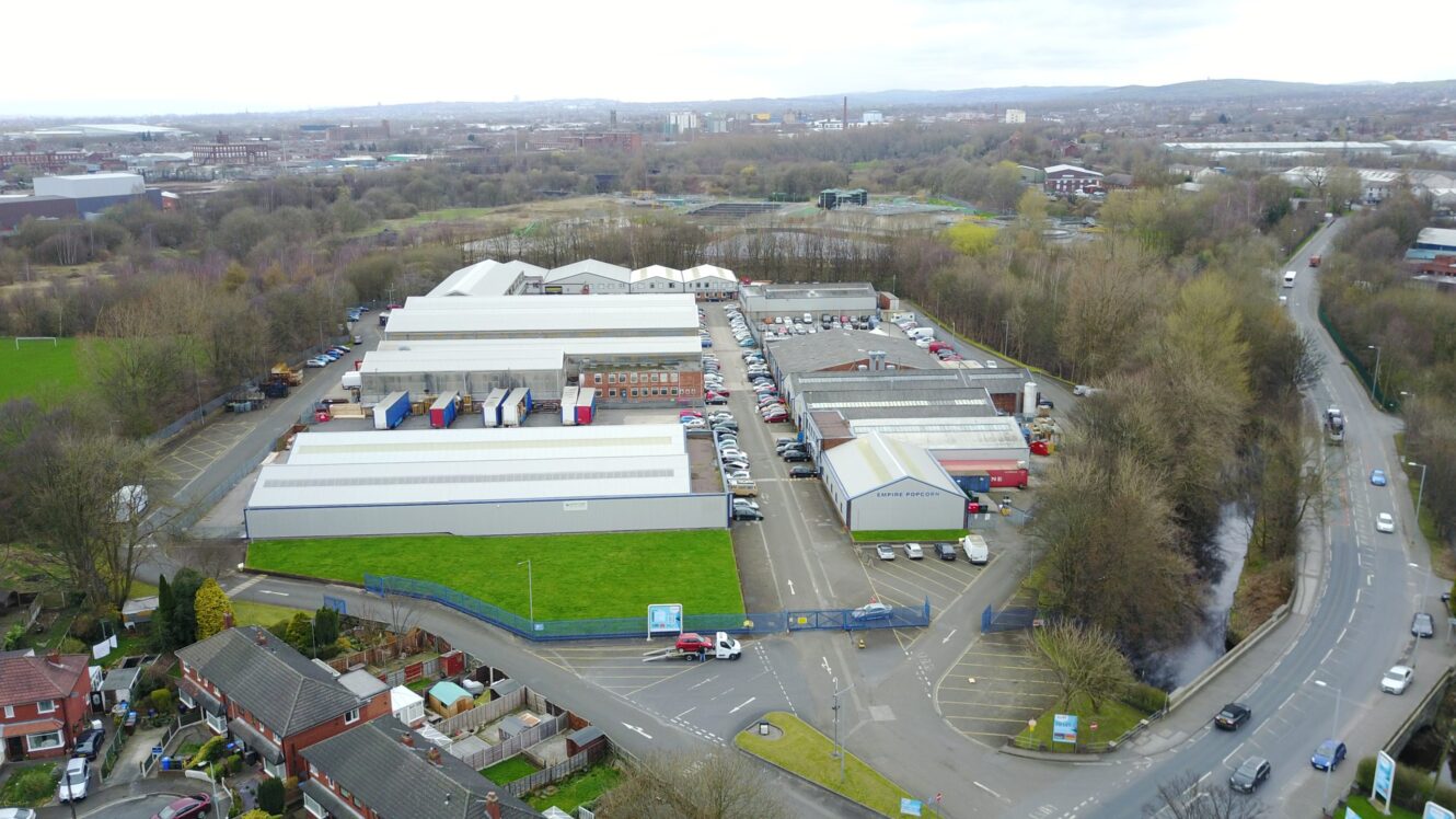 Aerial view of an industrial complex with several large warehouse buildings, parking areas, and surrounding roads, bordered by trees and residential houses.
