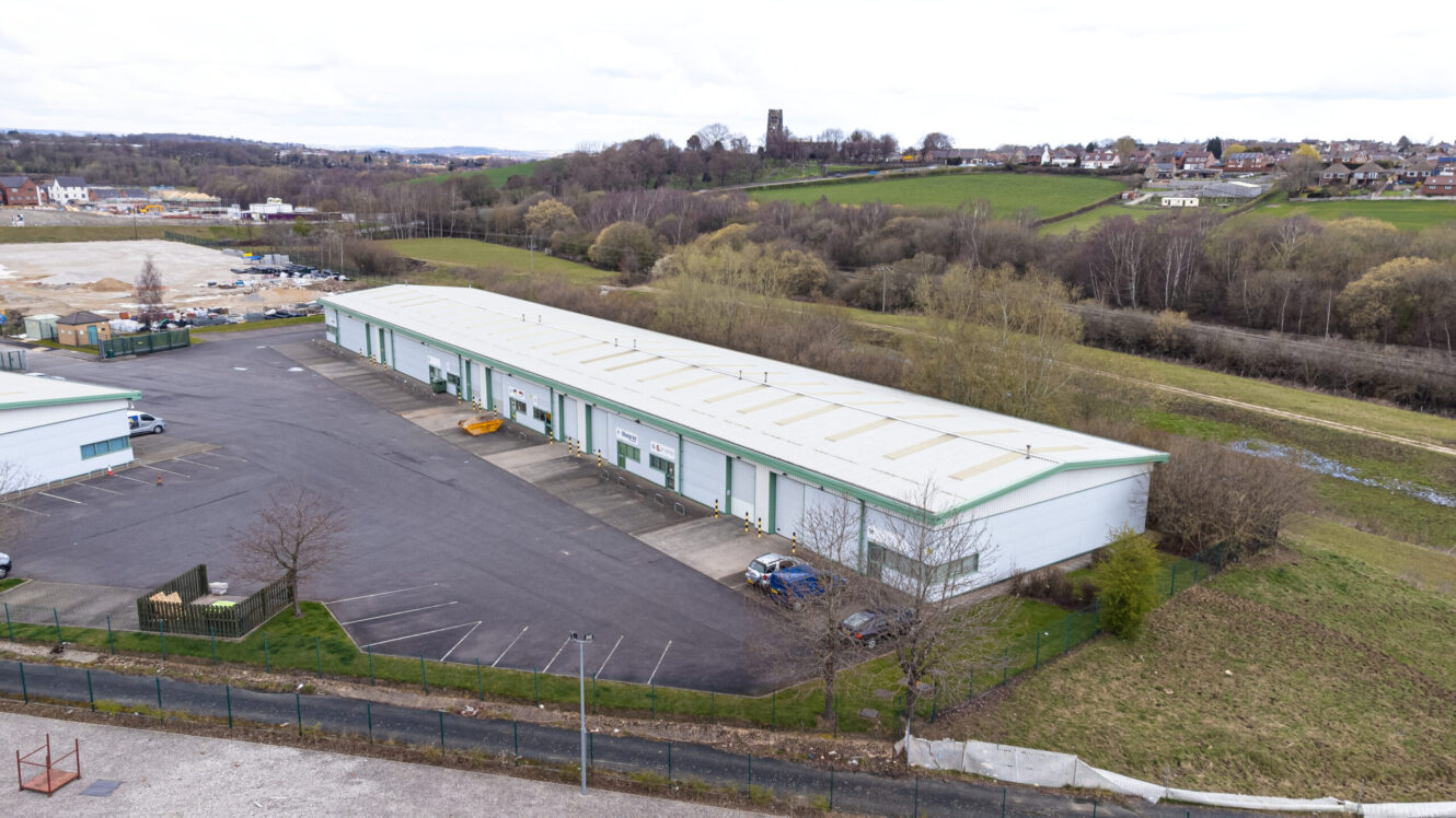 Aerial view of a large warehouse building with loading bays and parked vehicles, surrounded by an empty parking lot and a grassy landscape with trees and distant houses.