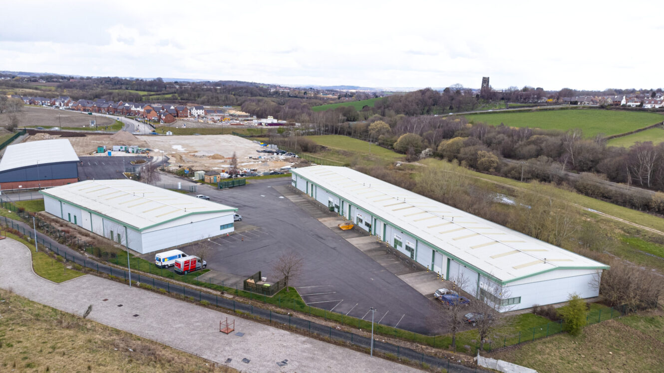 Aerial view of two large industrial warehouse buildings with adjacent parking, surrounded by fields and a small residential area in the distance.