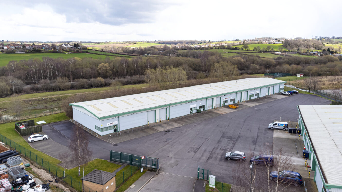 Aerial view of a single-story industrial warehouse building with several garage doors, surrounded by parked cars and greenery in the background.