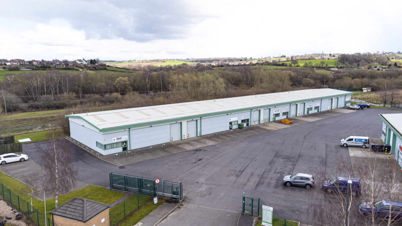 Aerial view of a long, single-story industrial warehouse with multiple garage doors and parked vehicles, surrounded by trees and open fields.