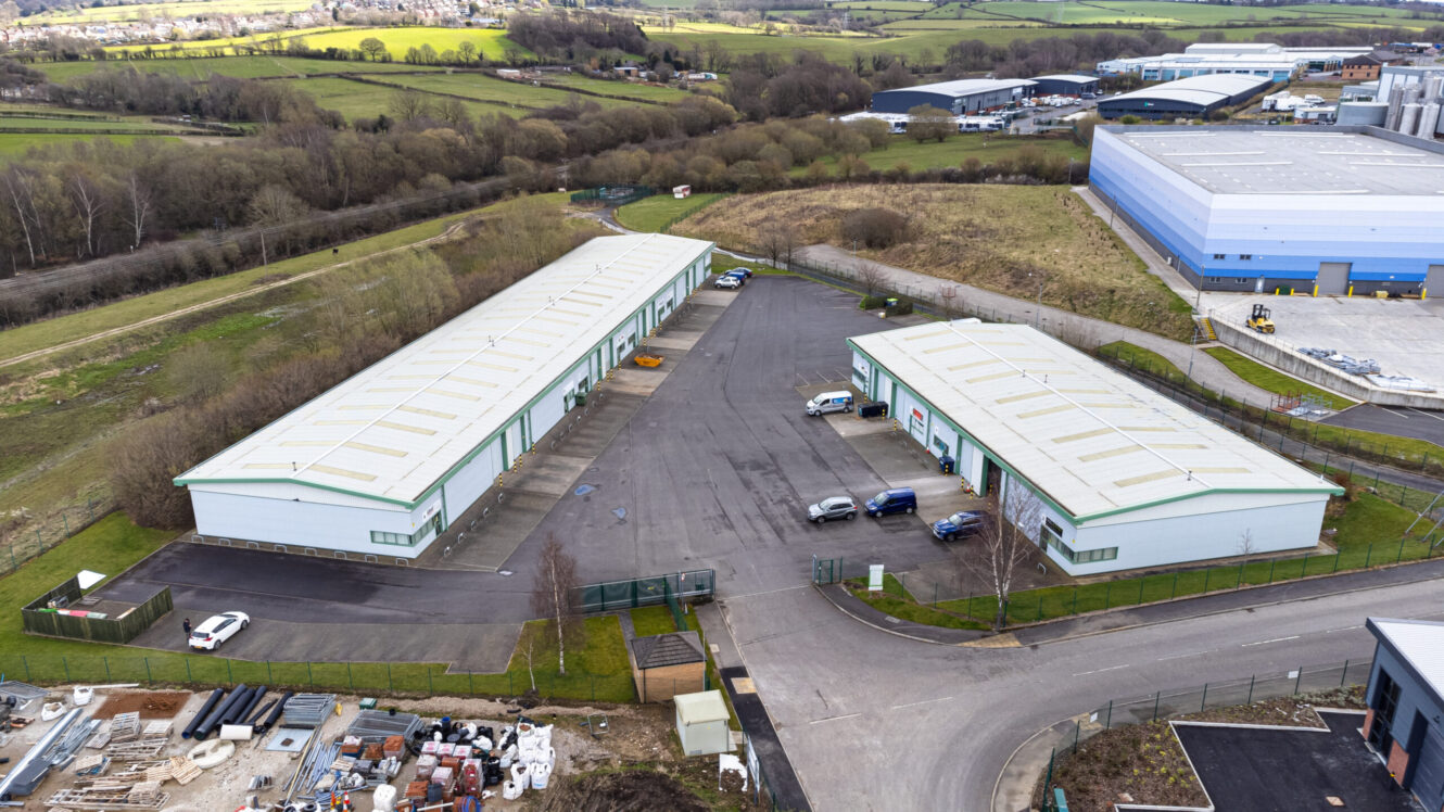 Aerial view of two large warehouse buildings with parking lots and several cars, surrounded by fencing and green landscape in an industrial area.