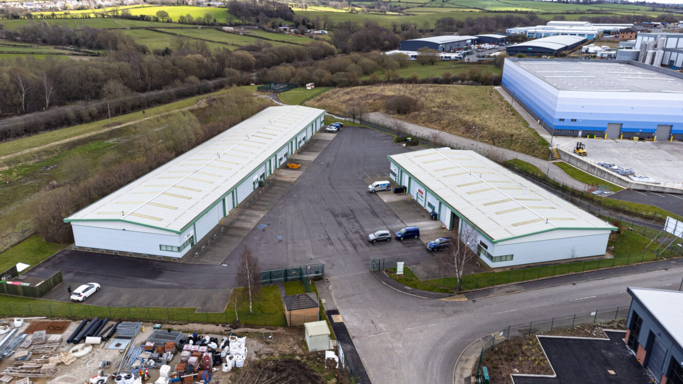 Aerial view of two long industrial warehouse buildings with parking lots, surrounded by roads, grassy areas, and other industrial facilities.