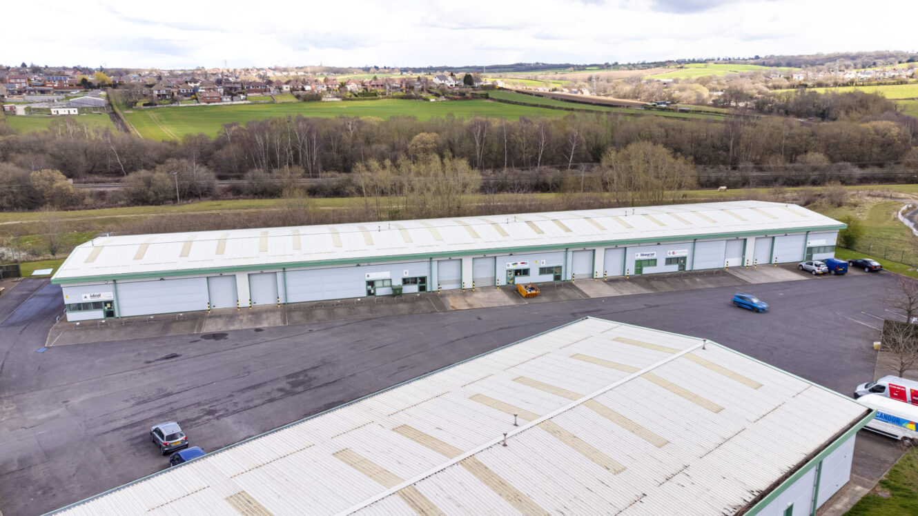 Aerial view of a long industrial warehouse with multiple units and roller doors, surrounded by parking spaces, grass fields, and a few parked vehicles.