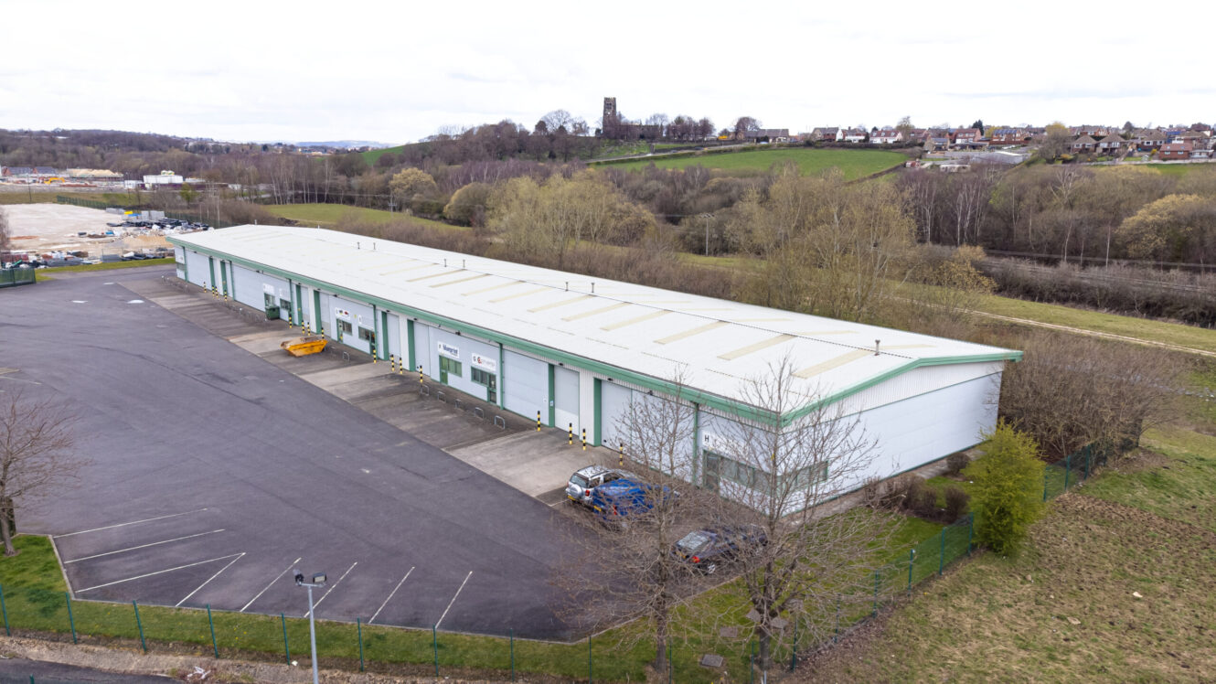 A large industrial warehouse with multiple loading bays is surrounded by a paved parking area and a grassy landscape, with a small town visible in the background.