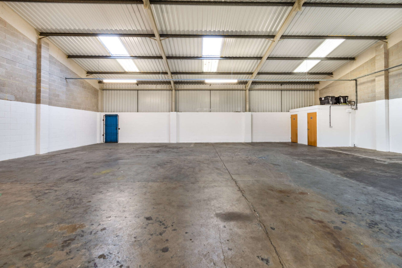 Empty industrial warehouse space with concrete floor, high ceiling, fluorescent lighting, exposed beams, a blue door on the left, and two brown doors on the right.