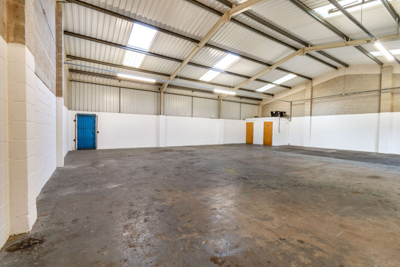 Empty industrial warehouse with concrete floor, white painted walls, metal roof, blue door on the left, and brown double doors on the right. Bright lighting from windows in ceiling.