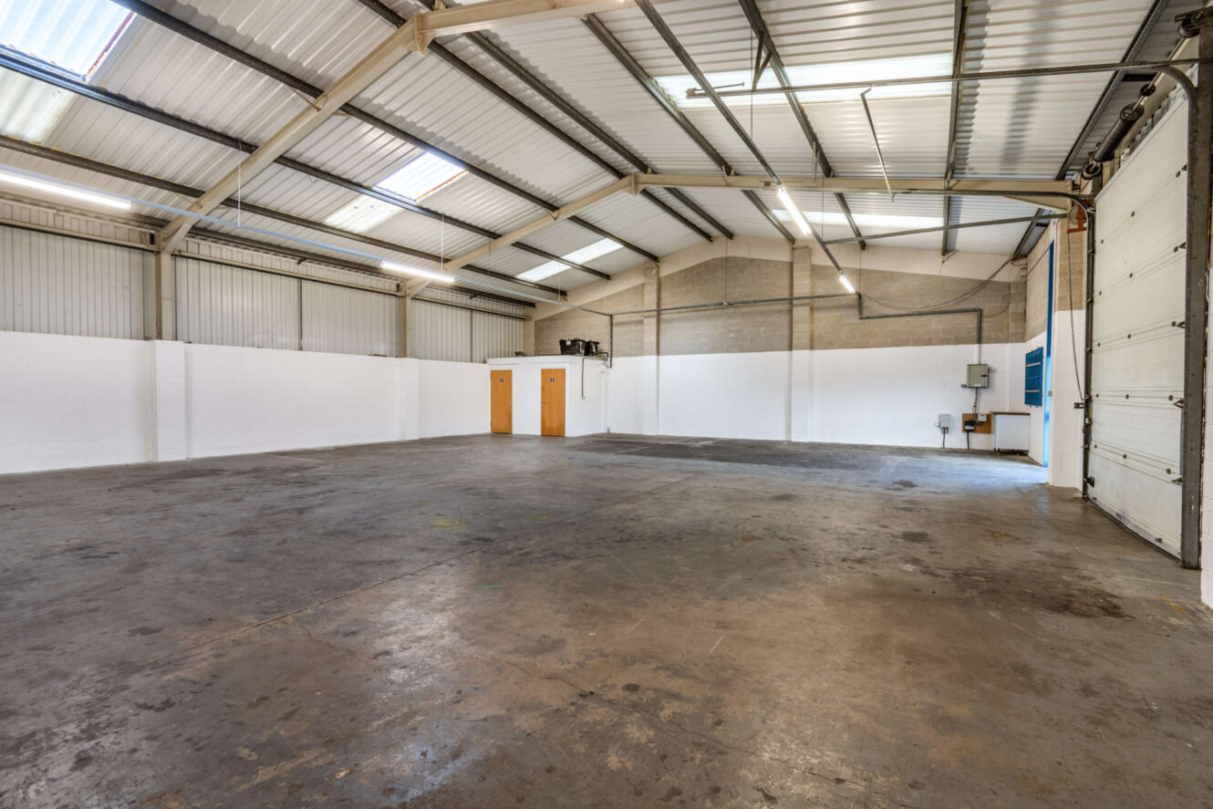 Interior of an empty industrial warehouse with concrete floor, white and gray walls, high ceiling, fluorescent lighting, and large rolling door.