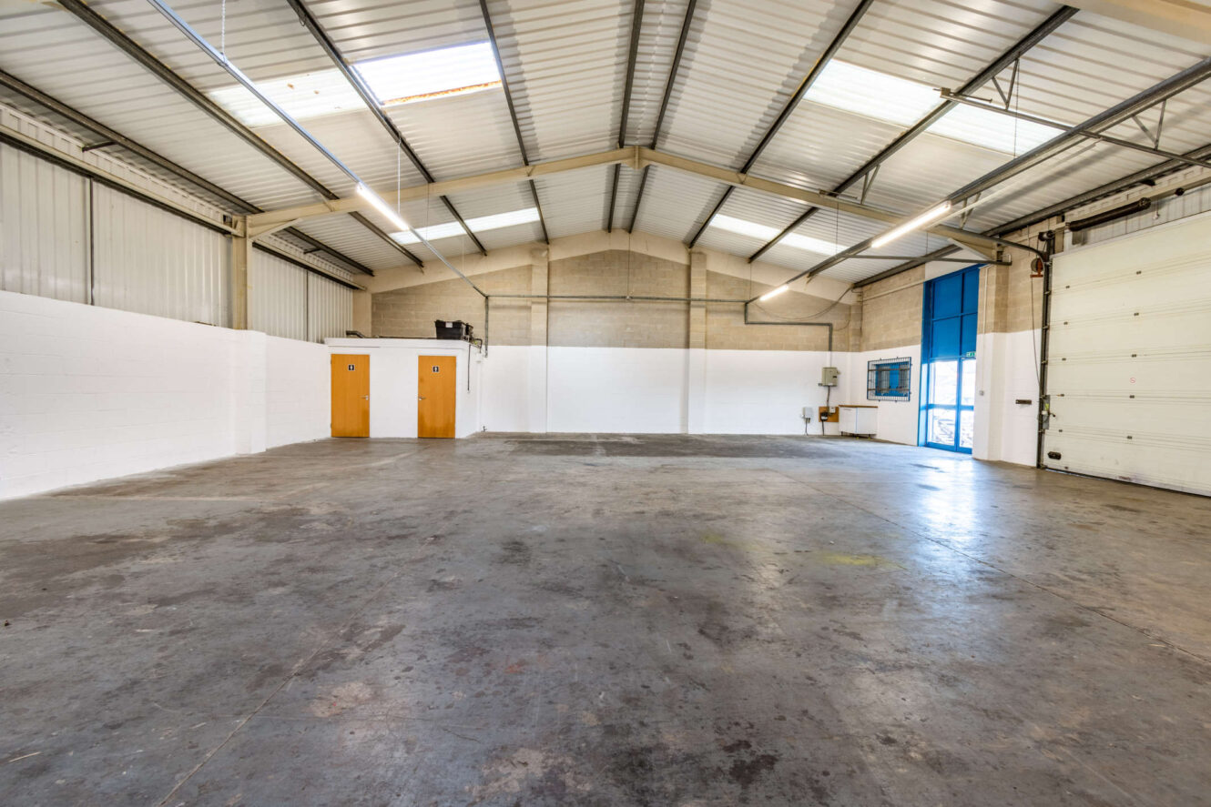 Empty industrial warehouse interior with concrete floor, high metal ceiling, two wooden doors, a large white garage door, and a blue entrance door.