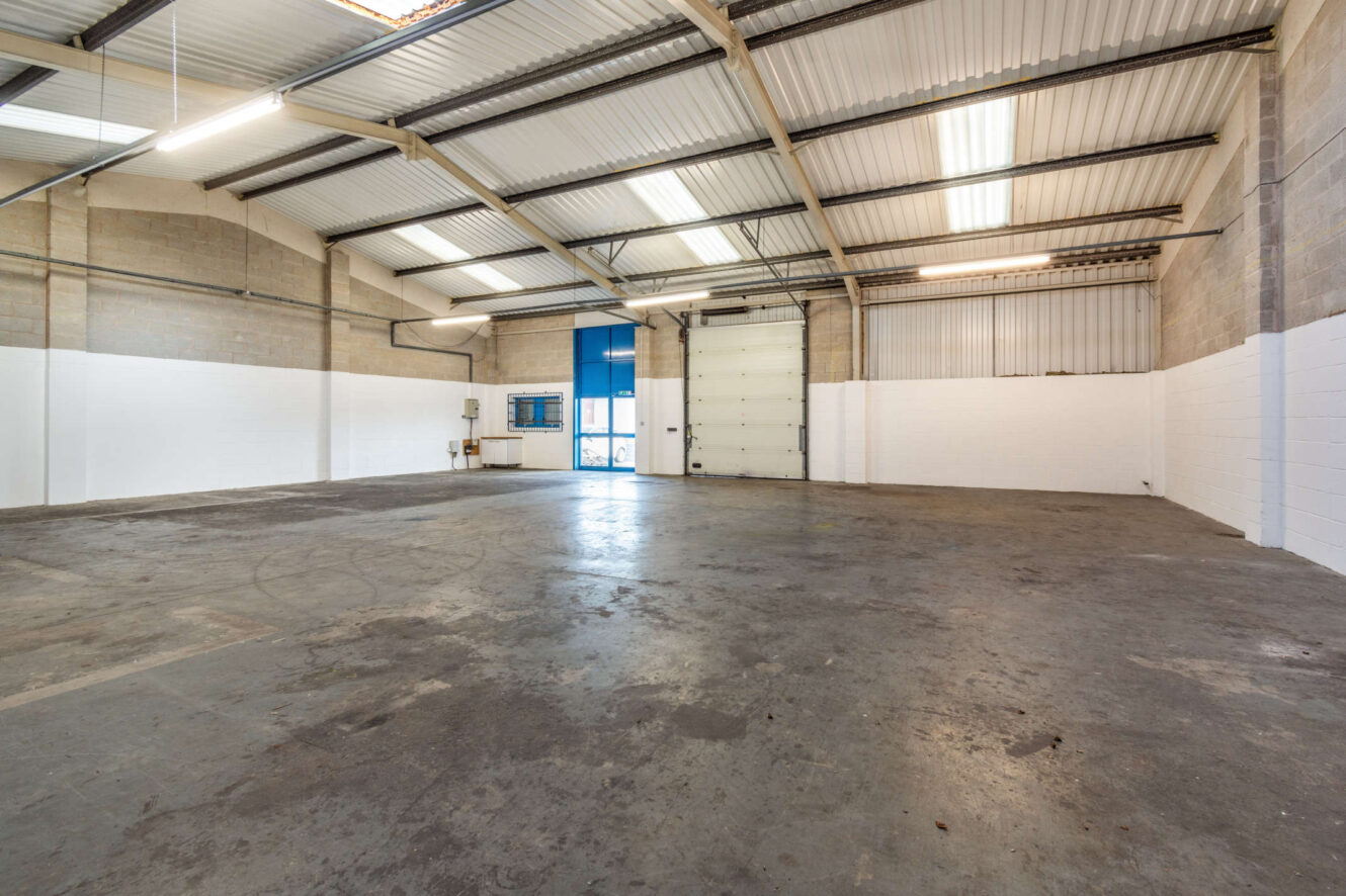 Empty industrial warehouse interior with concrete floor, exposed beams, white and beige walls, large roller door, fluorescent ceiling lights, and a blue window blind.