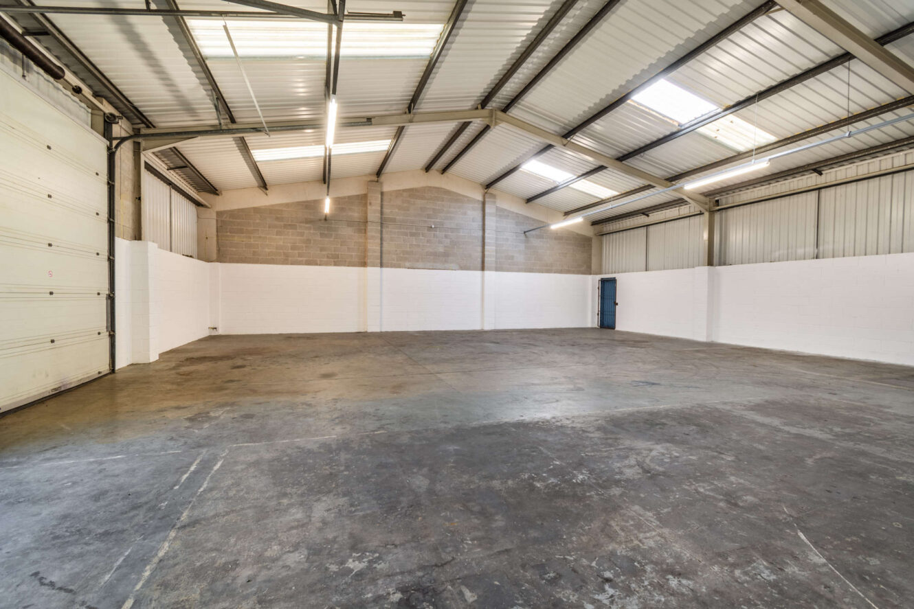 Empty industrial warehouse with concrete floor, white and brick walls, metal roof, fluorescent lighting, large garage door, and a blue entry door on the right side.