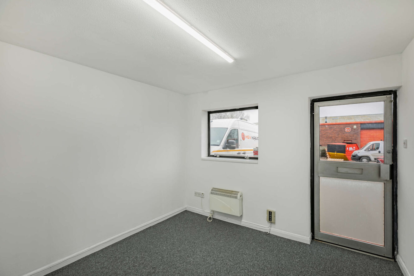 Small, empty office with white walls, grey carpet, a window and a door; a van and a red-brick building are visible outside.