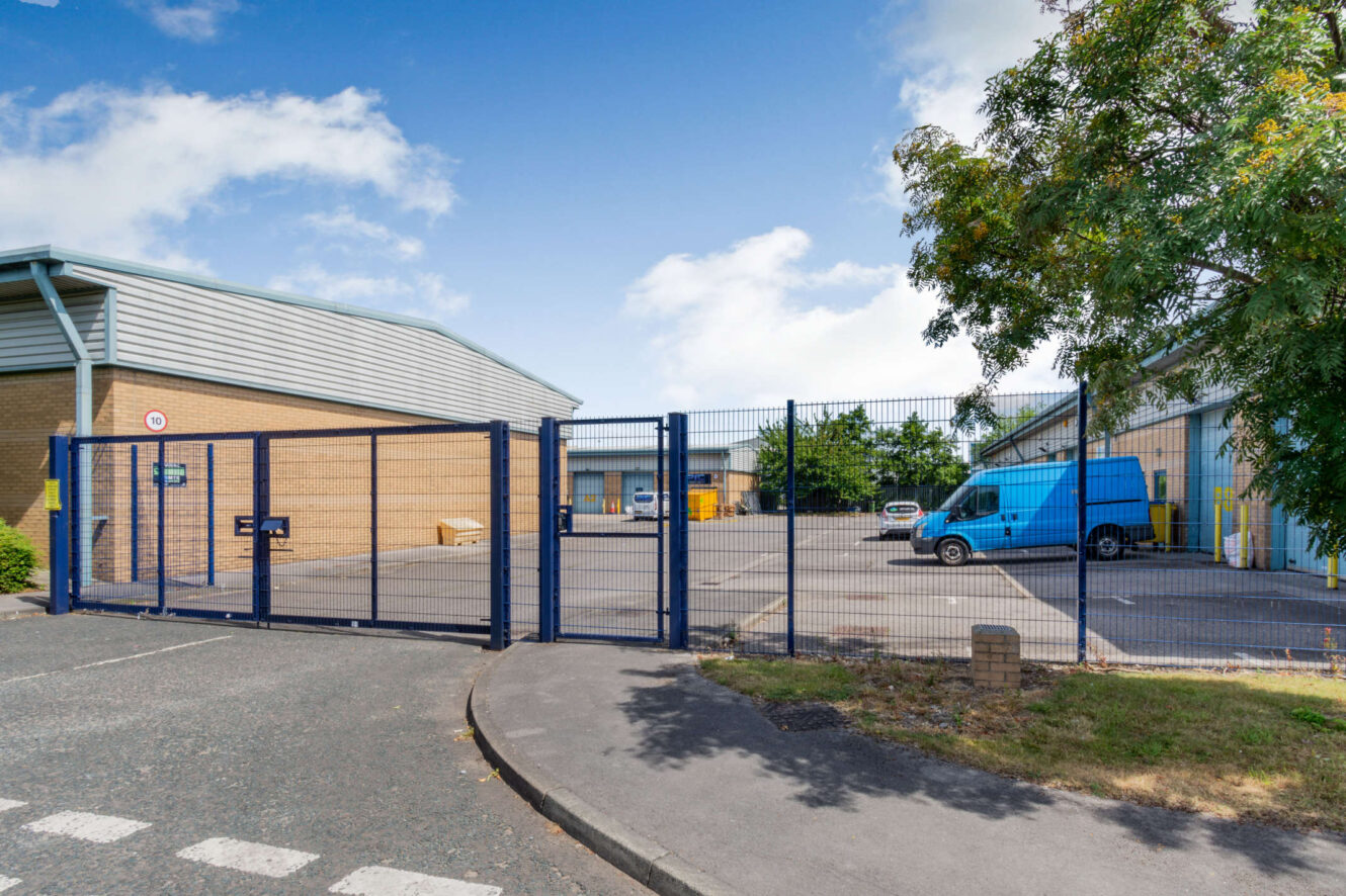 A gated entrance to an industrial or commercial complex with a blue van parked inside and buildings in the background on a clear, sunny day.