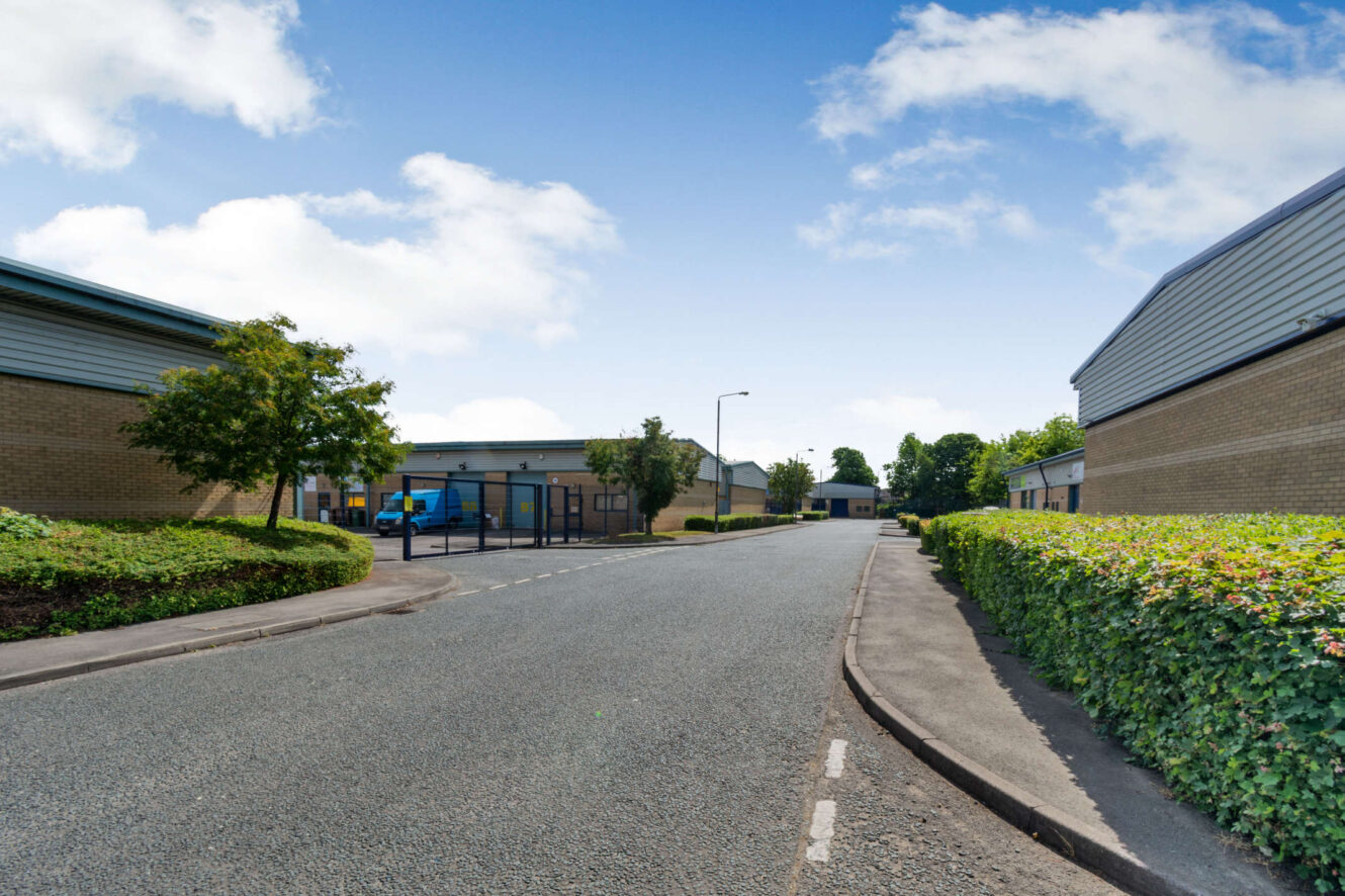 An empty industrial estate street with warehouses on both sides, trees, hedges, and a blue sky with scattered clouds.