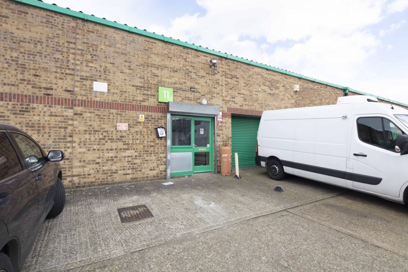 A brick industrial building with a green door and number 11 sign, flanked by a white van and a dark car, with a green roller shutter door partially visible.