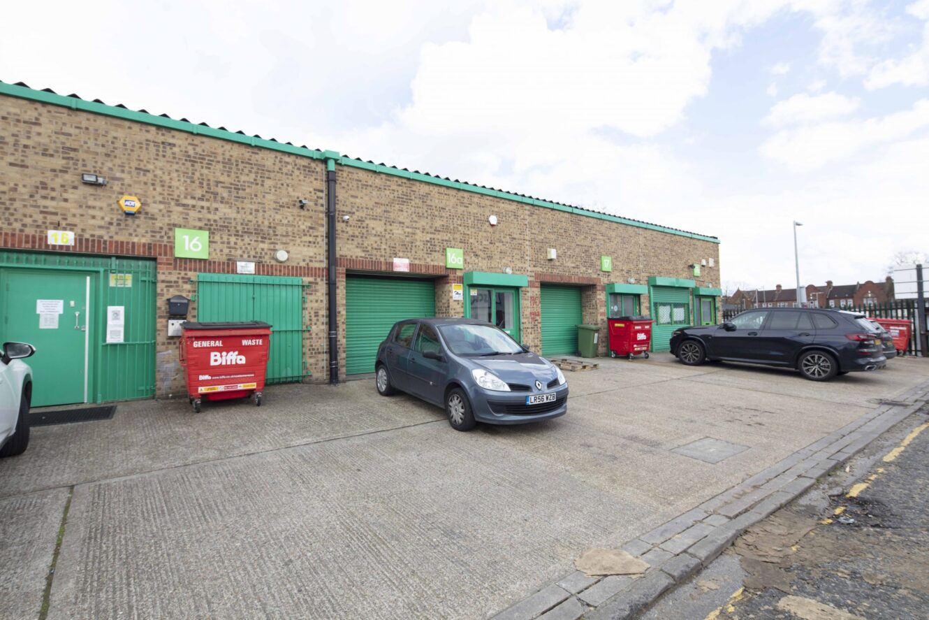 Parking area in front of brick industrial units with green doors, two parked cars, and red waste bins, on a cloudy day.