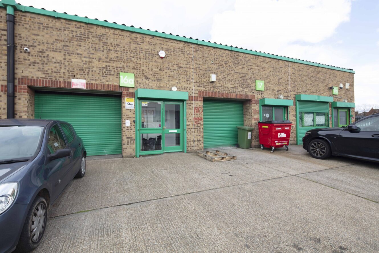 Two green roller shutter doors and a glass entrance on a brick industrial building, with two parked cars and a red waste bin outside.