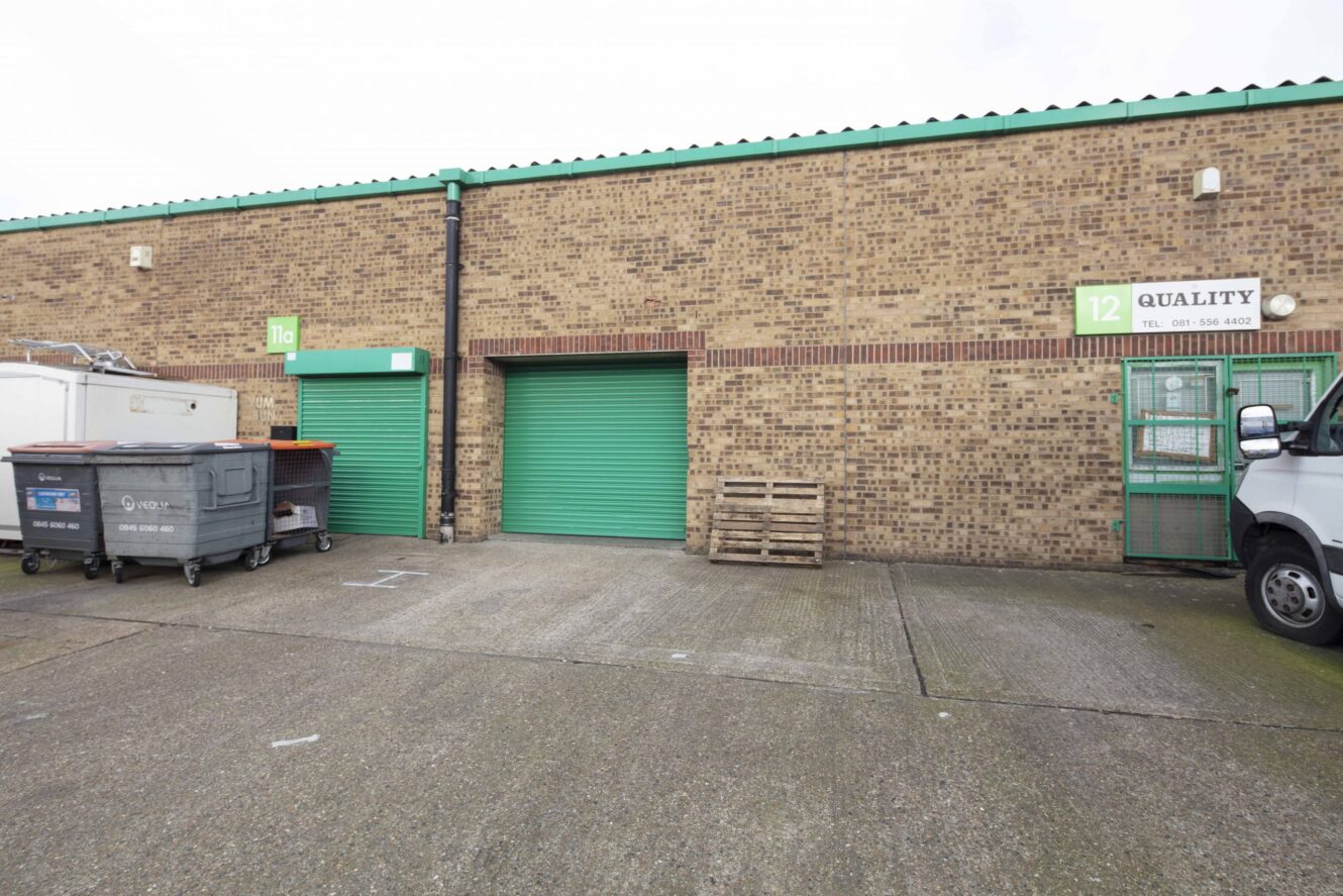 A brick industrial building with two green roller doors, a stack of pallets, large waste bins, and a sign labeled 12 QUALITY above a green door.