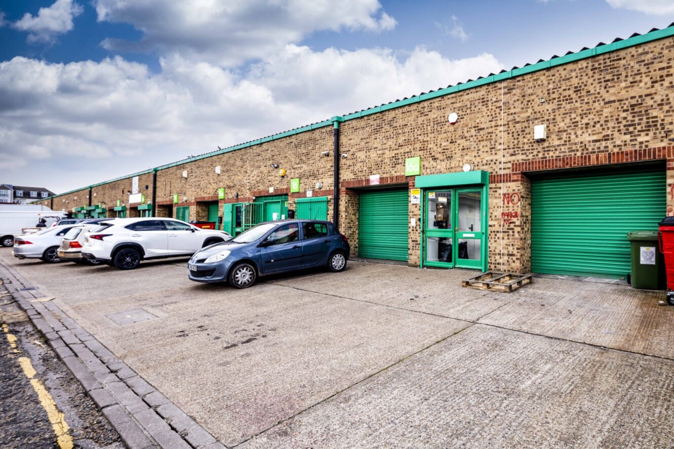 Row of industrial warehouse units with green doors and parked cars in front on a cloudy day.