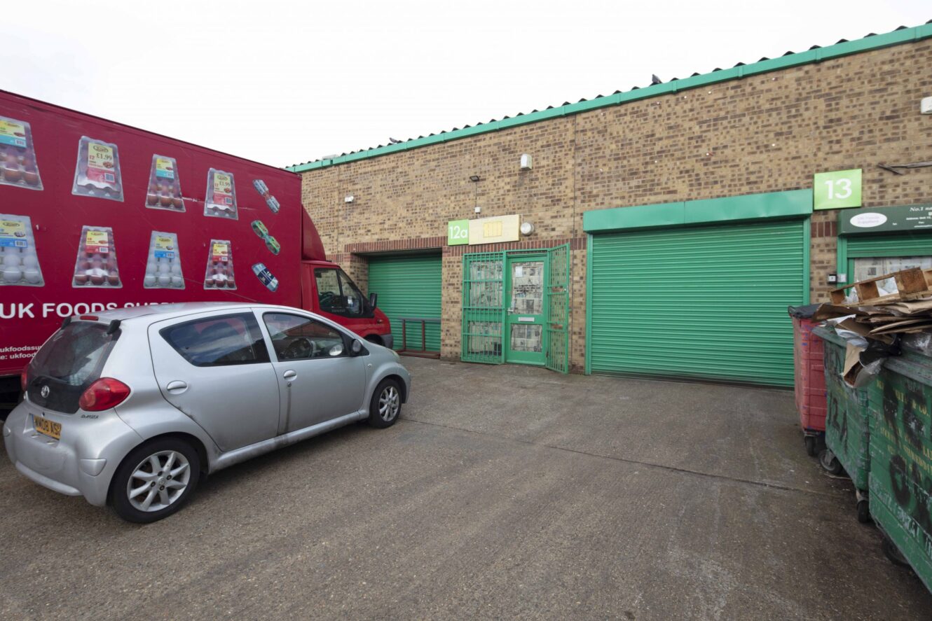 A small silver car and a red delivery truck are parked outside warehouse units with green shutters, numbered 12a and 13. A large dumpster is on the right side.