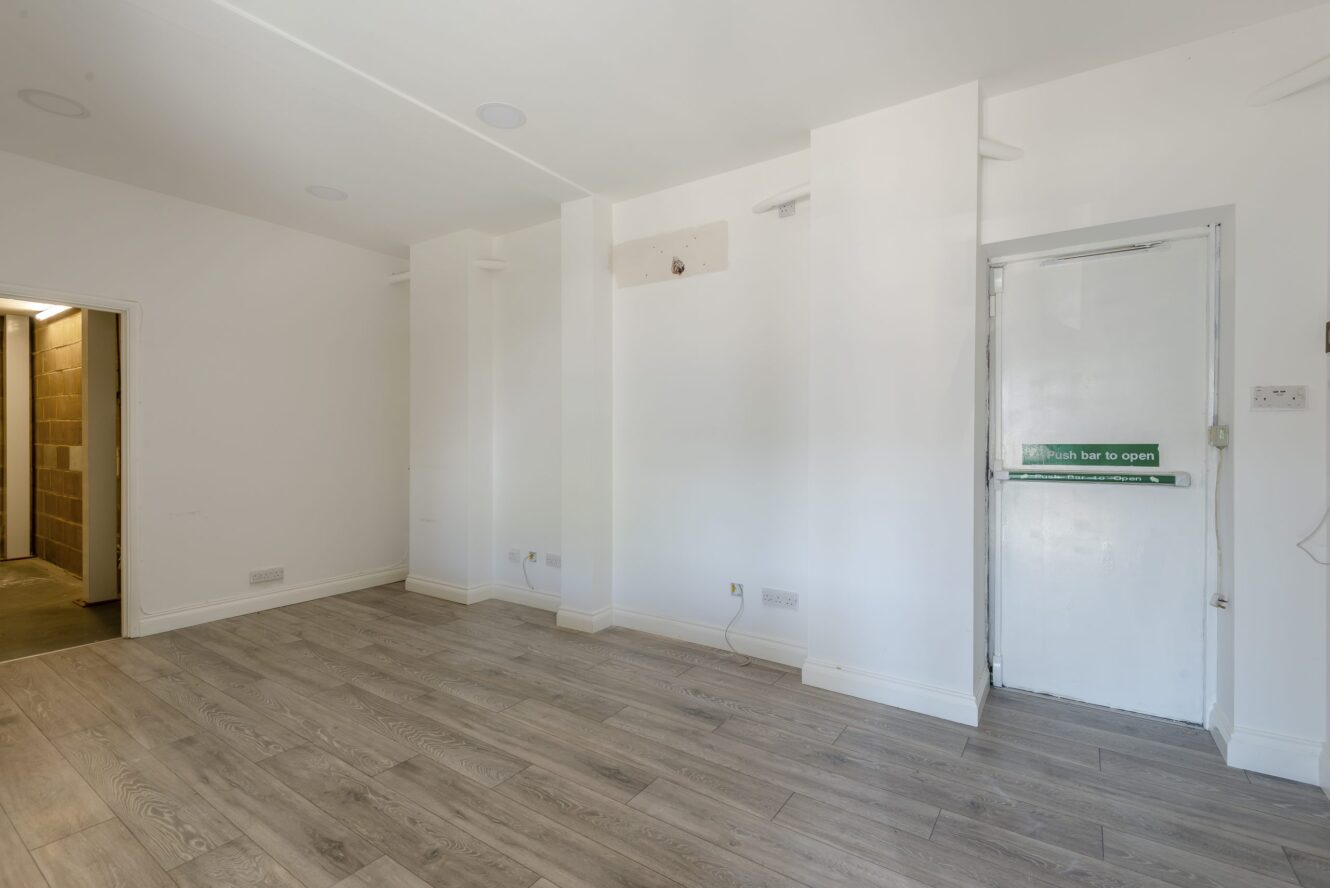 An empty room with light wood flooring, white walls, electrical outlets, a partially open door with a Push bar to open sign, and a doorway leading to another area.