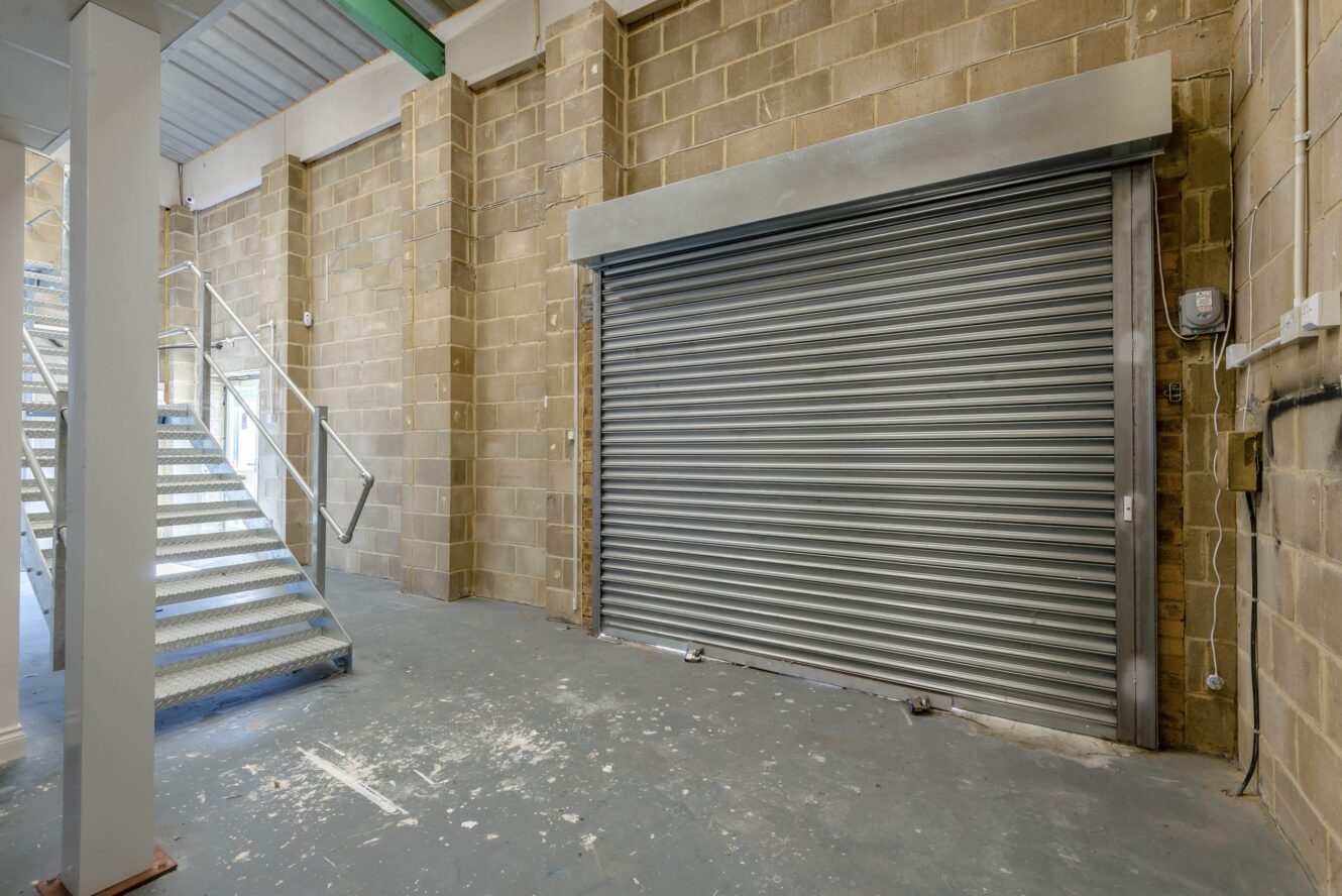 A metal roller shutter door in an industrial warehouse space with brick walls, a concrete floor, and a metal staircase.
