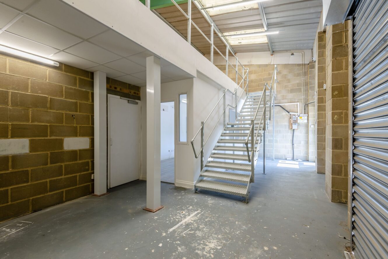 Interior of an industrial or warehouse space with concrete floor, brick walls, metal staircase, and a mezzanine level under fluorescent lighting.