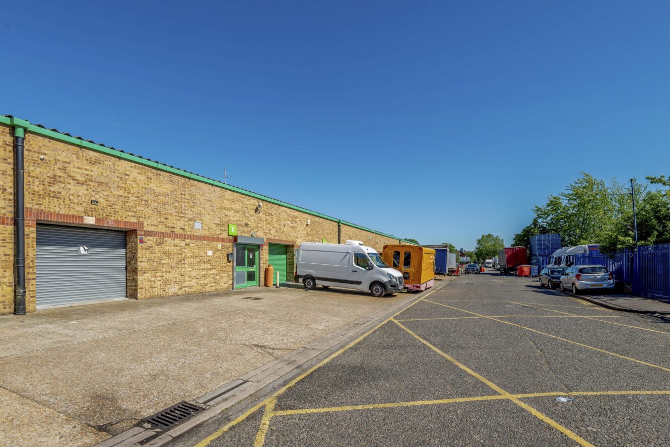 A light industrial warehouse with a yellow brick exterior, a van parked outside, and a clear blue sky overhead. Other vehicles are visible along the street.