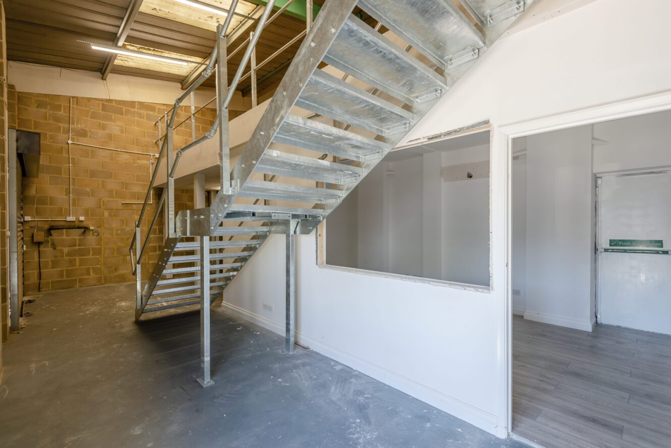 Industrial interior with a metal staircase, brick and white walls, a large window opening, and adjoining rooms with different flooring materials.