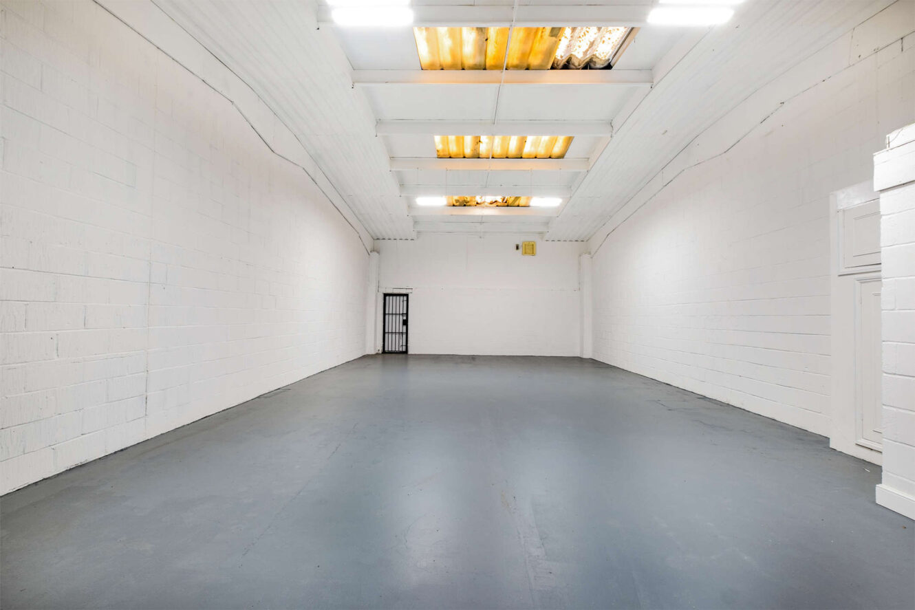 A large, empty industrial room with white brick walls, a gray concrete floor, and skylights in the ceiling. A metal door is visible at the far end.