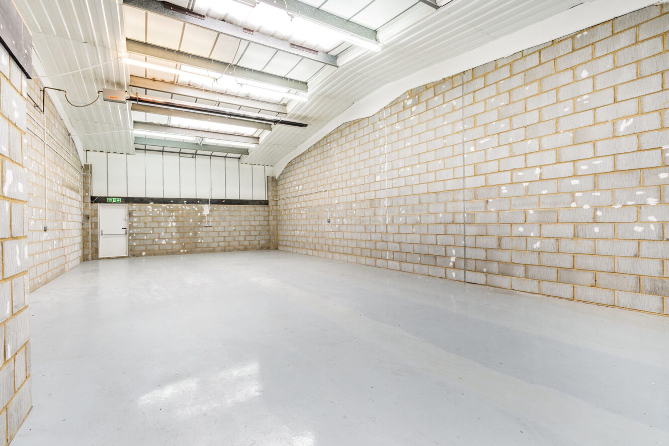 Empty industrial warehouse with white painted concrete floor, exposed block walls, and skylights in the ceiling. A white door and green exit sign are visible at the far end.
