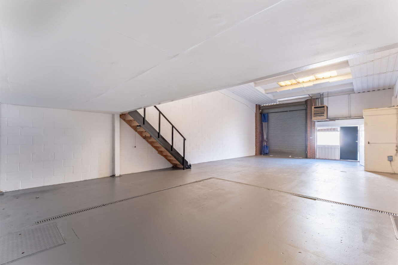 Empty industrial warehouse interior with white walls, grey floor, exposed beams, metal staircase, and a large roll-up garage door.