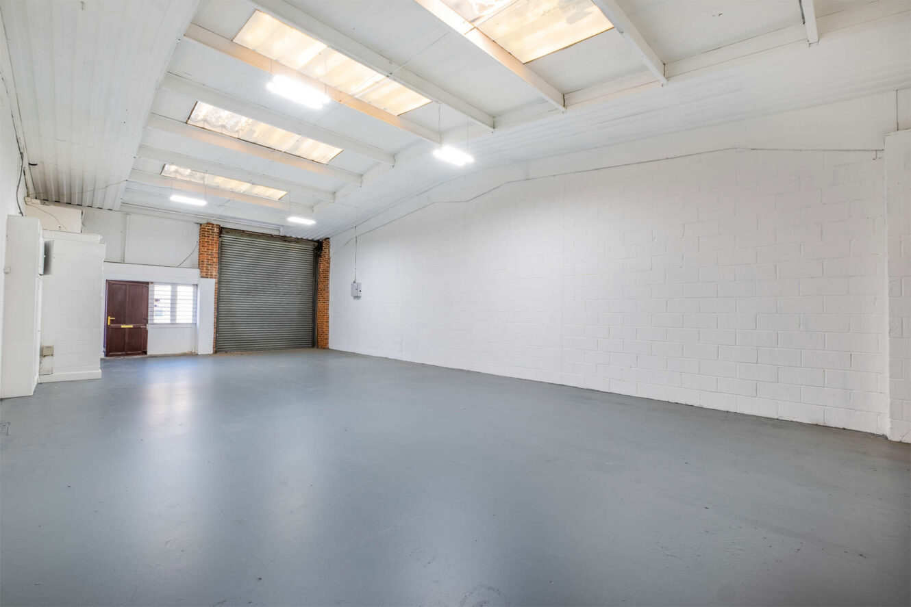 Empty industrial warehouse interior with white painted brick walls, a gray concrete floor, skylights, fluorescent lights, and a large roller door.